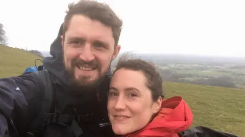 Aurelie Boas A man with brown hair and a brown beard and wearing a navy blue coat. A woman with brown hair and red coat. They are smiling. Behind them are green hills. The sky is grey and cloudy. 