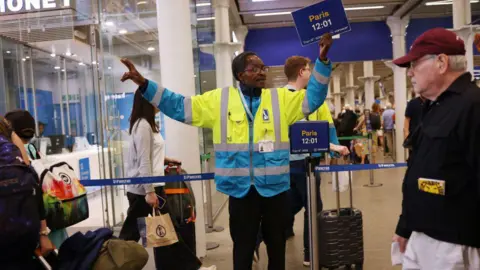 Getty Images Male member of staff in train station wearing a yellow and blue high-vis jacket, holding his arms out and holding a sign saying Paris 12:01
