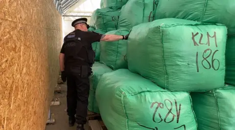 Essex Police A police officer in a room, touching a very large green bag, that looks to be full of clothing. He is wearing black, with a police hat on, his back is to the camera. 