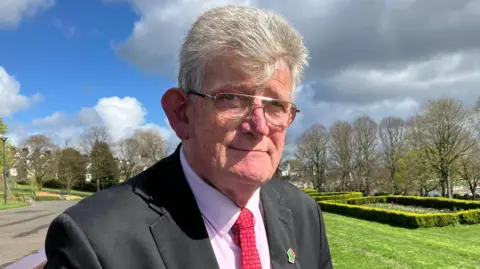 Jon McCourt wears a black suit jacket with a pink shirt underneath. He wears a red tie with a green black and red pin in the shape of a fist on his lapel. Behind him are green gardens and tree's. 