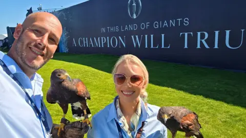Clear Skies Falconry A man and woman stand side by side, each dressed in a blue shirt and each holding a Harris hawk on their well protected arms. Both people are smiling. Both birds, which are a mix of shades of brown in colour, are motionless. A sign behind them showcases the Open championship golf tournament.