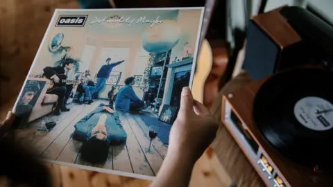 Caroline Briggs/BBC Two hands hold the album cover of Oasis' Definitely Maybe as the record plays on a nearby record player. The cover shows Liam Gallagher lying on the wooden floor of a living room, with a glass of what appears to be red wine next to him. Noel Gallagher sits on a sofa holding a guitar. Next to the sofa is a picture of Burt Bacharach. Three other band members are seen in various poses, one sits on the floor looking at a television, one sits on a chair and another stands in front of the window.