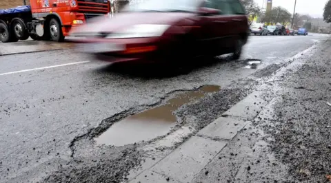 A red car driving close to a pothole filled with water