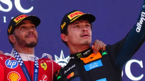 Lewis Hamilton and Lando Norris on the podium at the Barcelona Grand Prix on 14th June 2026. The winner, Hamilton, is in a red Ferrari racing suit covered in sponsorship logos. Norris, who came third, is in black for McLaren - also covered in sponsors.
