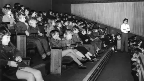 A black and white photo of children sat in rows at the cinema watching a film. There is an adult man standing up with his arms crossed, also watching the film. 