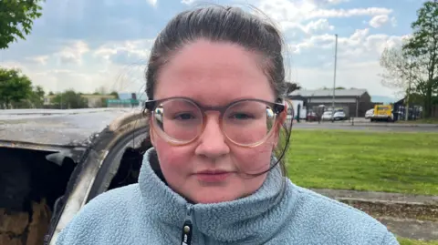A woman with black, pulled-back hair is standing beside the charred remains of a car. She is wearing black-rimmed glasses and a light blue fleece.
She is standing near a large grassy area, with several cars visible in the background.