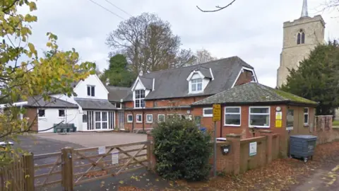 A primary school with a yard in to the left and old two-storey brick buildings to the right. There is a white building with a triangular upper floor in the background, and a brick wall in the foreground with a wooden gate across the entrance. There is a church to the right.
