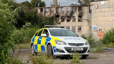 A police car in front of the Saxondale site which has large, black burn marks on the walls.
