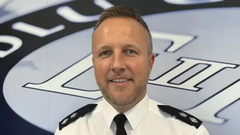 A man with short brown hair and brown and grey facial hair. He is wearing a white shirt with black police patches on the shoulders. He wears a black tie and is standing in front of a large background with a gwent police logo on it