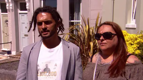 A man wearing a T shirt and light coloured jacket next to a woman with an off-the-shoulder dress and wearing dark glasses.