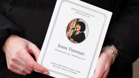 Hands holding an order of service which has a photo of Jeane Freeman on the front