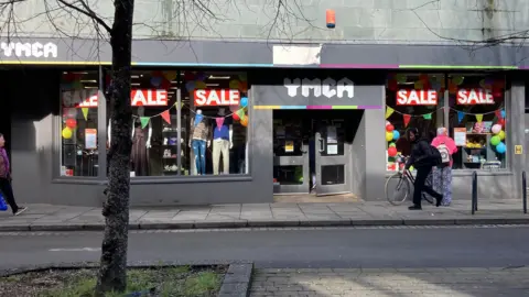 BBC A photo showing a YMCA charity shop in Gloucester with a front door, windows and a tree outside