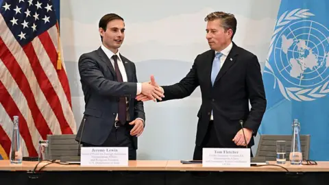AFP via Getty Images Two suited men stand in front of an American flag and a UN flag, shaking hands. They are US Under Secretary of State for Foreign Assistance, Humanitarian Affairs and Religious Freedom Jeremy Lewin and United Nations Under-Secretary-General for Humanitarian Affairs Tom Fletcher.