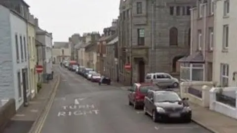 Google A view looking down High Street in Port St Mary against the flow of traffic. It is a narrow street with tall buildings on either side and cars parked on the right.