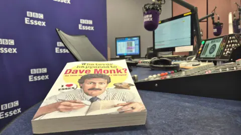 Stuart Woodward/BBC A book entitled "Whatever happened to Dave Monk?" sits on a desk in a radio studio. There are microphones, headphones and mixing desks in the background, as well as a large purple background with several white BBC Essex logos on it.
