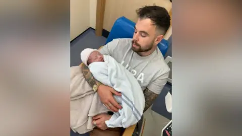 Dom Edington Dom Edington wearing a t shirt holds his new born baby daughter who is wrapped in a towel and wears a knitted hat.