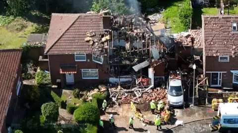 Aerial view of two semi-detached homes. The one on the right has been obliterated, with a massive pile of rubble, broken roof rafters and badly damaged internal walls visible. Houses either side have missing roof tiles. There are a number of people in hi-vis jackets standing in front.