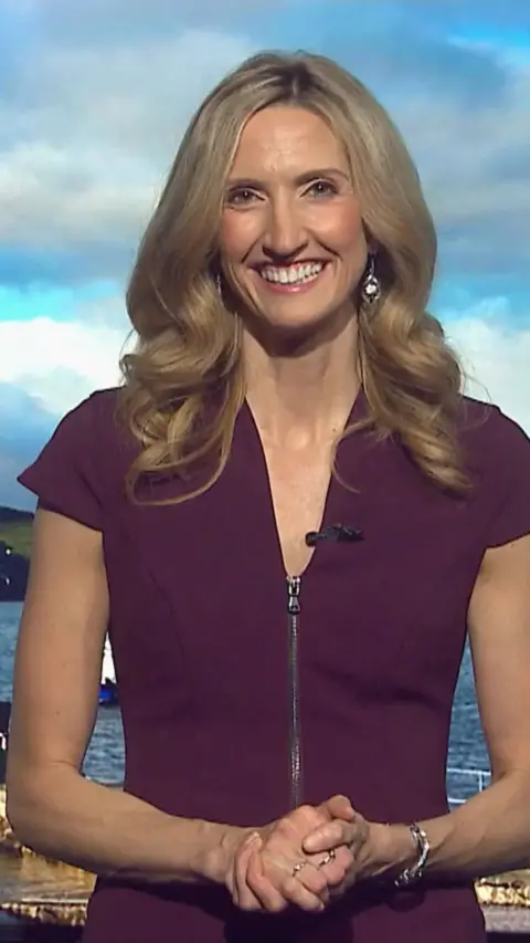 A smiling woman with long blonde hair, wears a burgundy short-sleeve dress, behind her is a picture of clouds