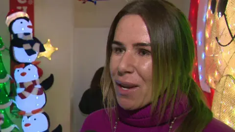 Rhiannon Canavan has long brown hair and is wearing a purple polar neck jumper and a beaded necklace. She is stood in front of an illuminated display of penguins and a Christmas tree