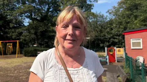 A woman with blonde hair, tied back with a fringe, stands in a children's playground in front of swings and a zip line. She is wearing a white top with the strap of a brown bag across her shoulder.