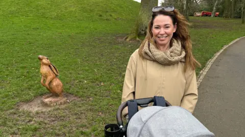 Katie Alexander, a woman with long, light brown hair, smiles at the camera in Wallace Park in Lisburn. She is pushing her daughter's grey and black buggy which is partially visible in the photo. Katie is wearing a beige coat, a matching woollen scarf and she has large sunglasses on top of her head. There is a small wooden statue of a hare to her left and a large tree behind her.