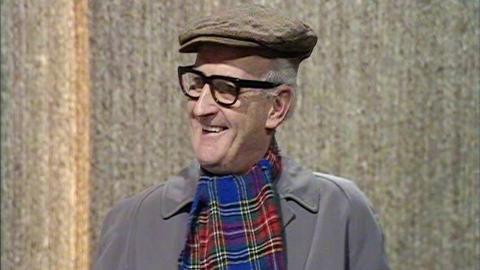 Walter McCorrisken in the studio. He's wearing a grey coat, tartan scarf, flat cap and thick black glasses. He's smiling.