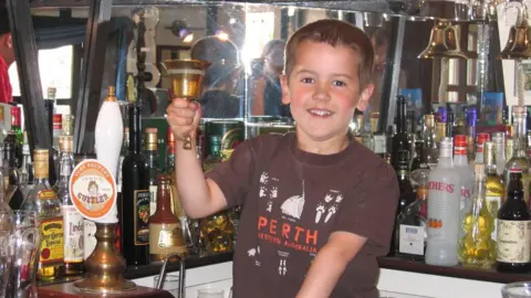 David Hull A child stands behind a bar counter, holding a small hand bell and surrounded by rows of bottles and drink dispensers. The counter is crowded with various glass bottles, labels, and tap handles, with a mirrored backdrop reflecting the setup. The scene is brightly lit, showing a casual indoor setting with many colourful containers and bar accessories.