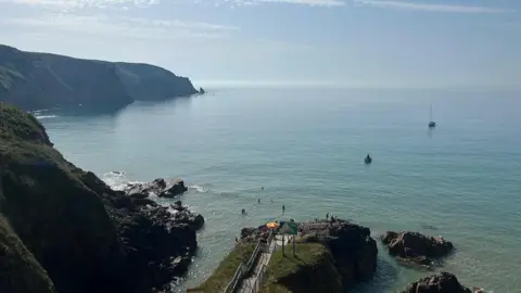 Steps lead down to the sea with headlands to the left. The sky is hazy and a small yacht can be seen offshore.