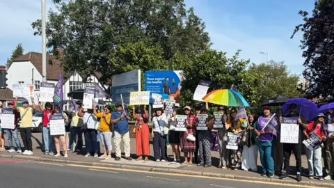 Unison UNISON members and supporters stand along a roadside holding union flags, placards, and umbrellas during a public demonstration. Many of the signs display messages such as “Fair Pay Now". The protesters form a long line beside a busy road under clear blue skies, creating a visible and organised presence. Purple UNISON branding is prominent throughout the gathering, highlighting calls for improved pay and support for workers.