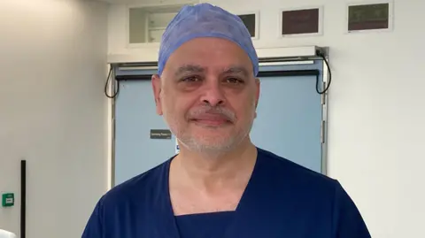 A surgeon is looking into the camera. He's wearing navy scrubs and a light blue covering on his head.