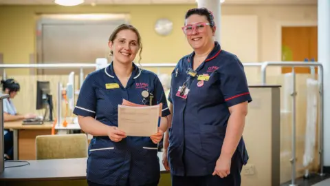 Mark Howe/St Luke's Two women are stood in a hospice in navy blue nursing uniforms and smiling at the camera. The woman on the left has light brown hair tied back and the woman on the right has black hair tied back and bright pink framed glasses