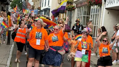 Smiling crowds of people dance down a narrow street 