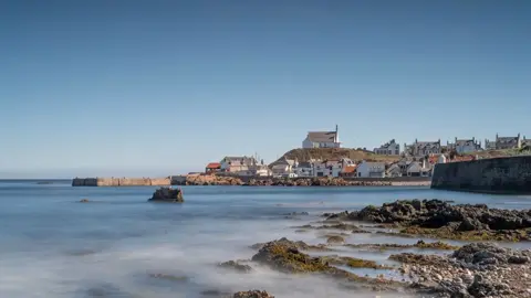 John Fotheringham A seaside village in the background with bright blue water and rocks along the coastline. The houses are white with orange and brown roofs. 