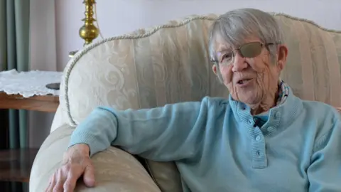 A picture of Eileen smiling at the camera. She is sat on a cream colour sofa and is wearing a blue shirt. She has glasses.