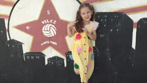 Nicolle Loughlin A young girl is standing against a mural painted on a wall. The mural features a large red star with the text “VELEŽ MOSTAR” and a ball graphic inside it, along with stylised city buildings in black and red rays radiating from the star. The child is wearing a yellow dress decorated with colourful circular patches and white trainers with pink details.