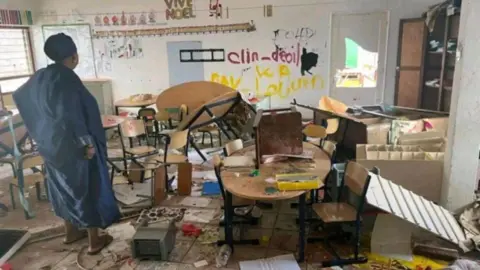 Getty Images A damaged classroom in Mayotte shows tables overturned and children's art on the floor. A woman looks at the scene - with a "Vive Noel" sign still visible on one of the walls.