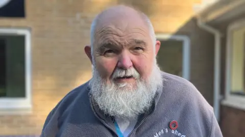 Brian McCluskey, who has a silver beard, is wearing a grey zip-up sweater with a Men's Shed logo on his left crest