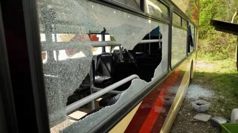 A coach with smashed windows. There is smashed glass on the grass outside.

