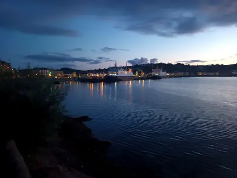 Lynn Martin A waterfront during early nightfall, with calm water, two large illuminated vessels, and a town stretching along the shoreline.