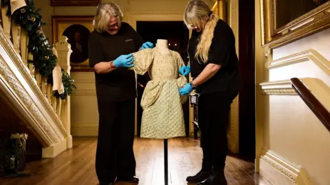 David Lindsay Two women dressed in black tending to a child's dress on a clothes stand. They are stood in the hall of a grand house.