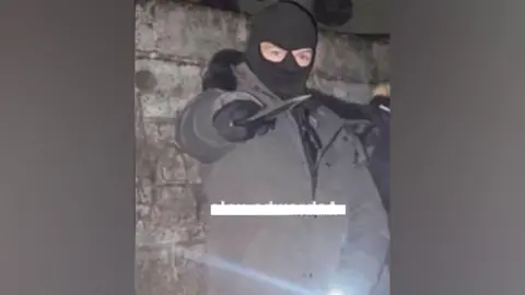 Counter Terrorism Policing Wales Alex Hutton wears a balaclava and poses with a machete
