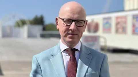Patrick Hurley has a bald head, dark spectacles, a white shirt, maroon tie and sky blue jacket,