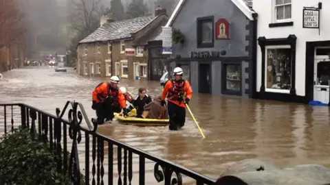 The flooded high street in Whalley, full of brown, knee high water. In the middle are people on a yellow raft being pulled through the water by men in orange jackets and white hard hats
