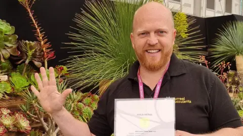 A mid shot of Daniel Michael. He is standing with his hand open to represent the five times his nursery has won the award. He is holding a certificate and is dressed in a black polo shirt.