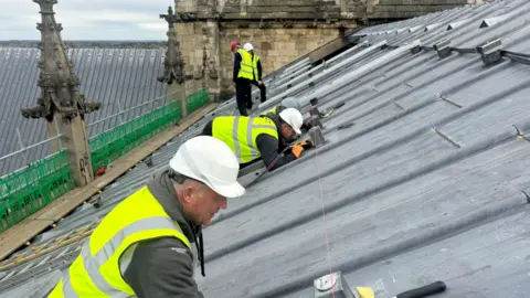 BBC/ Emily Johnson Five workers in helmets and hi-vis jackets crouched on the slanted roof of York Minster