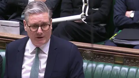 Prime Minister Sir Keir Starmer with grey hair and black framed glasses wearing a pale shirt, grey tie and navy suit speaking during Prime Minister's Questions in the House of Commons.