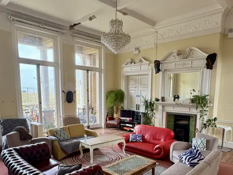 Katrina Woolley A large living room painted white with a chandelier hanging from the ceiling, a fireplace. 