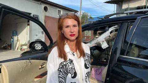 BBC / Imogen Anderson Olena Kryvchun, a woman with red hair and glasses perched on her head, wearing a white striped shirt and bright red lipstick, stands next to her car. A smashed windscreen, collapsed roof and debris-torn interior can be seen.