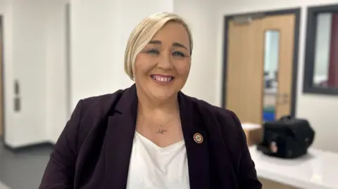 Fiona Kane is wearing a purple blazer over a white top. She has a small silver chain around her neck. The has short blonde hair and a badge on her blazer lapel. Behind her are white walls and a brown door. 