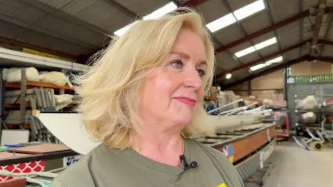 Image shows Eleanor Fitzgerald, a volunteer for Ukrainian Action. She has a blonde bob. She is looking off to the left of the picture. Behind her is the four dragon boats and one rowing boat which are being donated to Ukraine. She has a gold necklace on and a khaki green t-shirt.